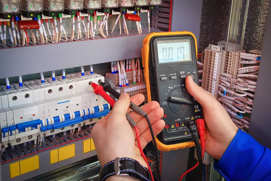 Manos con un polímetro digital y sus puntas de prueba midiendo en un cuadro eléctrico con interruptores