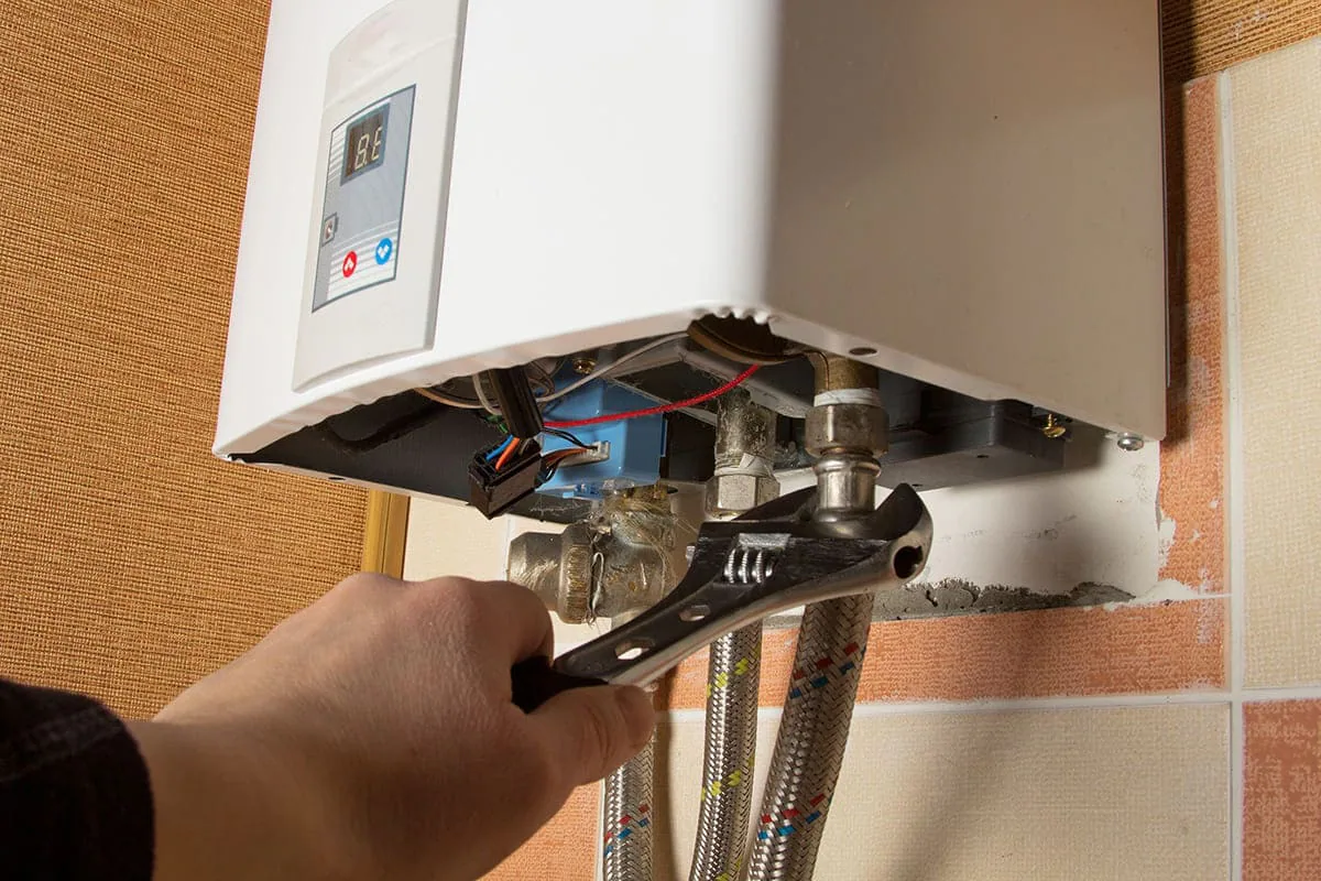 Mano que ajusta con una llave inglesa una conexión situada bajo un calentador de agua abierto con pantalla digital, imagen ilustrativa de reparación de calentadores de gas en Elche