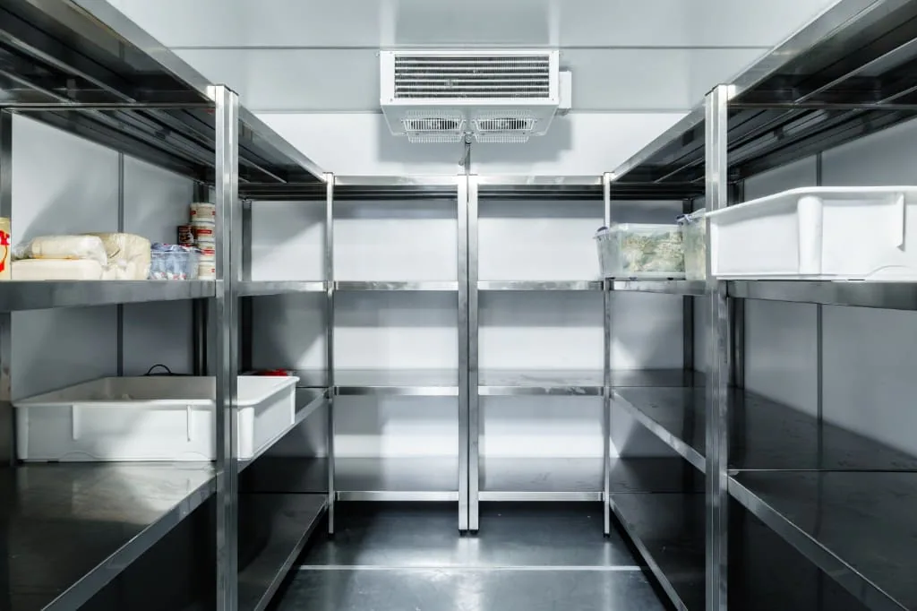 Interior de una cámara frigorífica con estanterías de acero inoxidable, recipientes de alimentos y evaporador al fondo