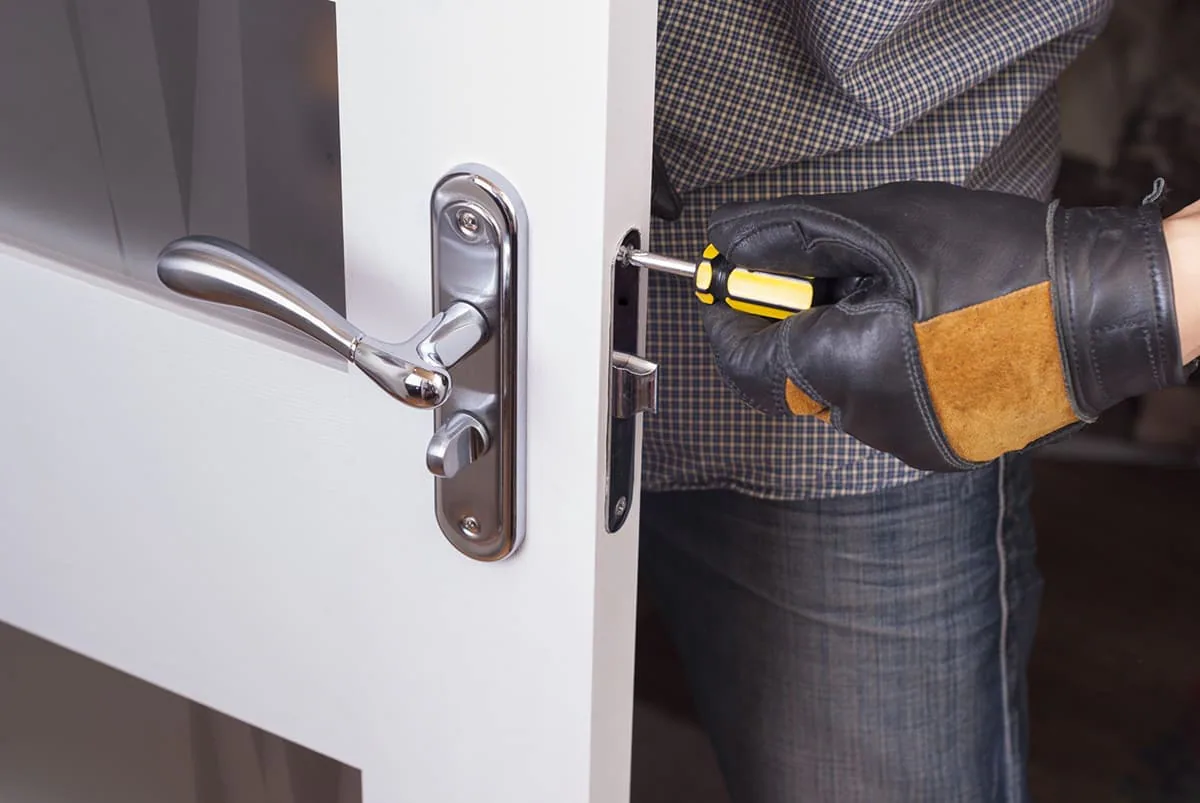 Mano con guante de cuero atornillando el frente de una cerradura en el canto de una puerta blanca