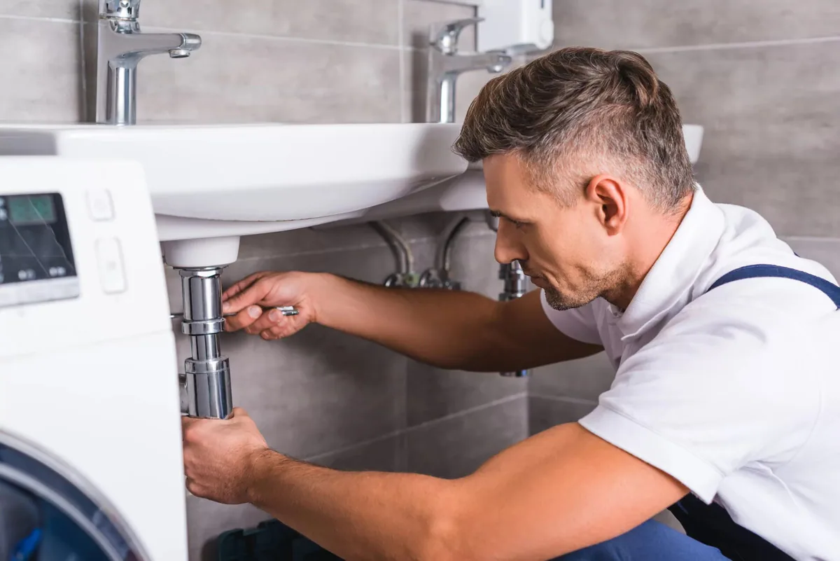 Operario desmontando el sifón cromado de un lavabo en un baño junto a una lavadora