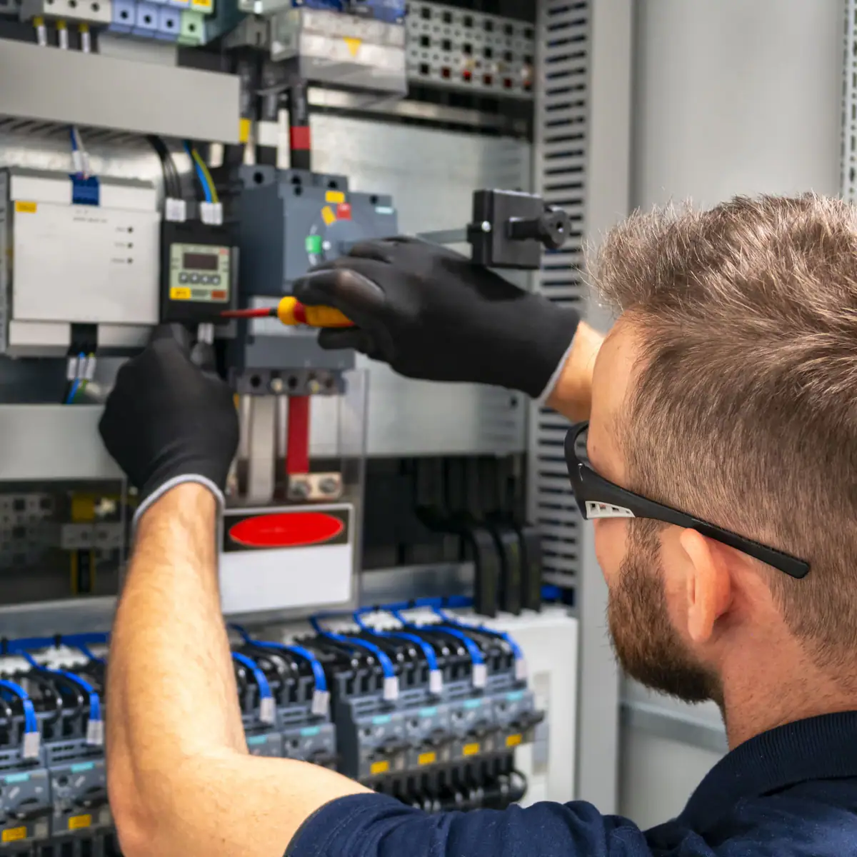Técnico con guantes negros y gafas de seguridad trabajando con destornillador en un armario eléctrico de gran tamaño