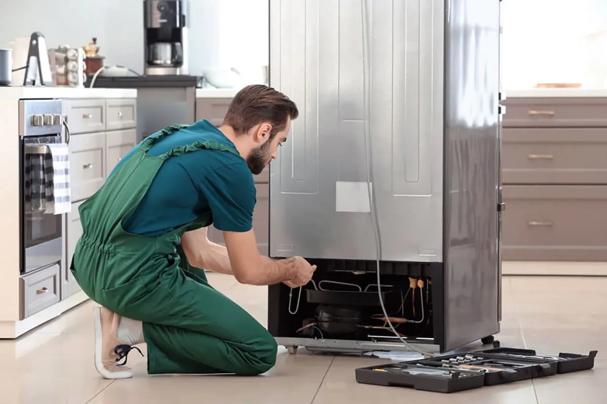 Técnico con peto verde arrodillado revisando el compresor trasero de un frigorífico, con caja de herramientas abierta, imagen ilustrativa de reparación de neveras en Tenerife