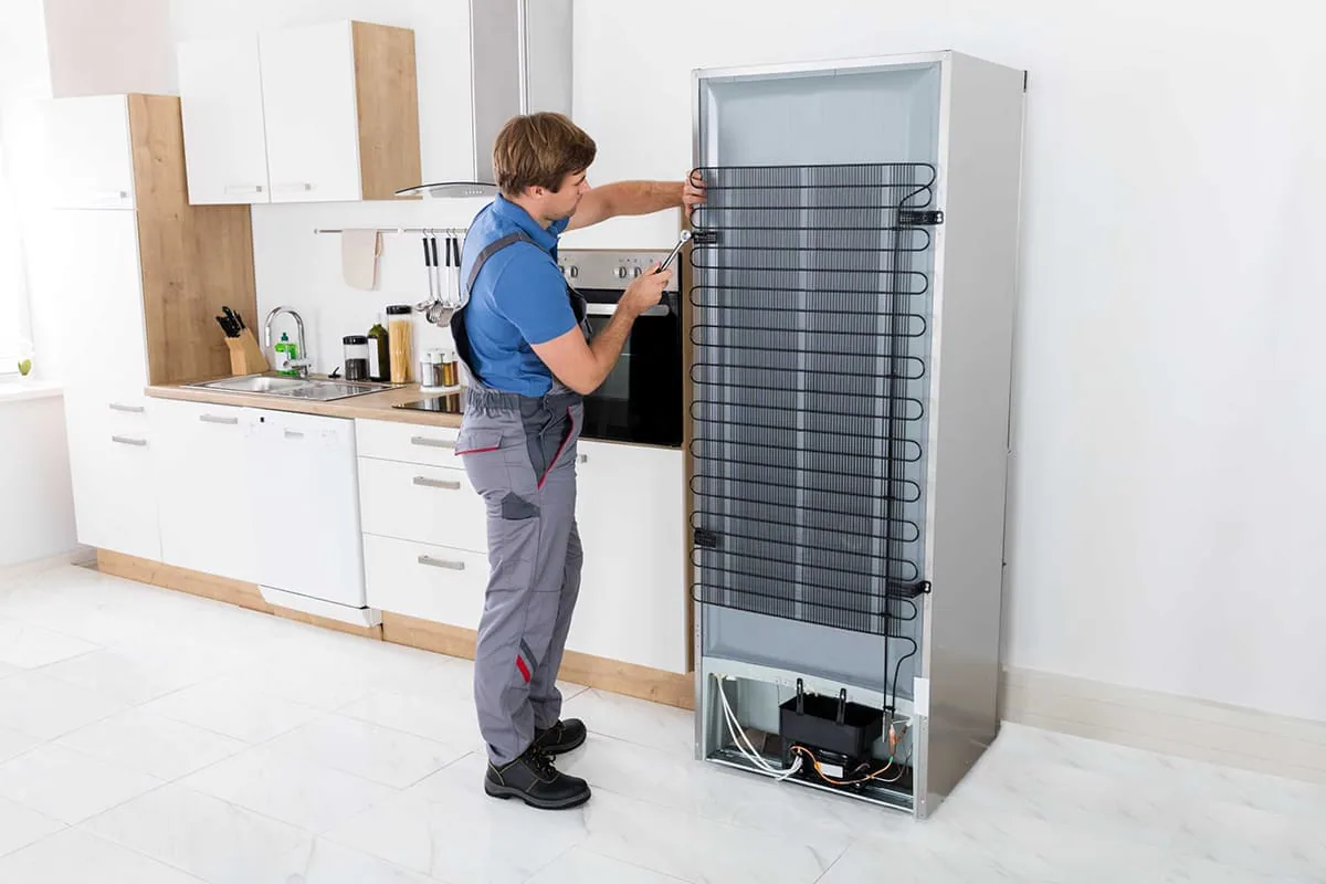 Técnico revisando con una llave el condensador trasero de un frigorífico apartado de la pared en una cocina