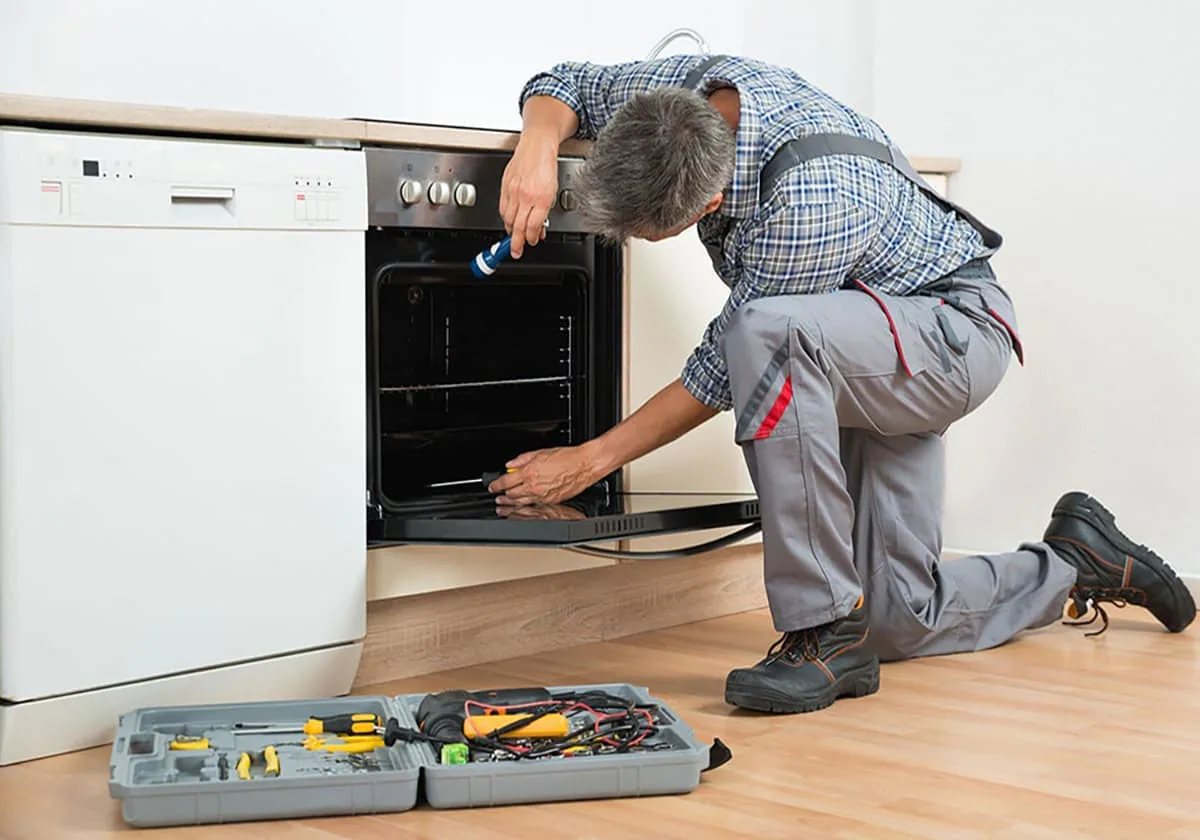 Técnico arrodillado que revisa con una linterna el interior de un horno empotrado junto a un maletín de herramientas
