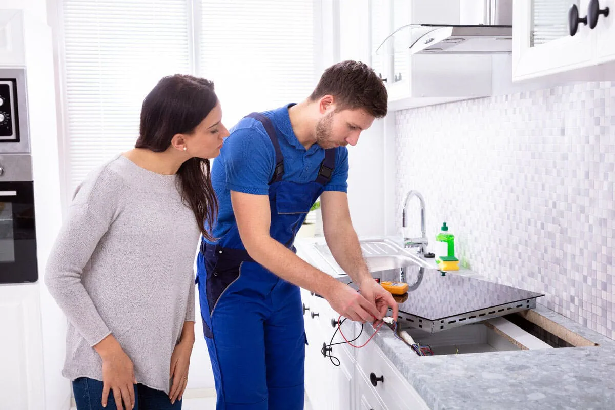Técnico con peto azul revisando los cables de conexión de una placa de cocina mientras una mujer observa, imagen ilustrativa de reparación de placas de inducción en Elche