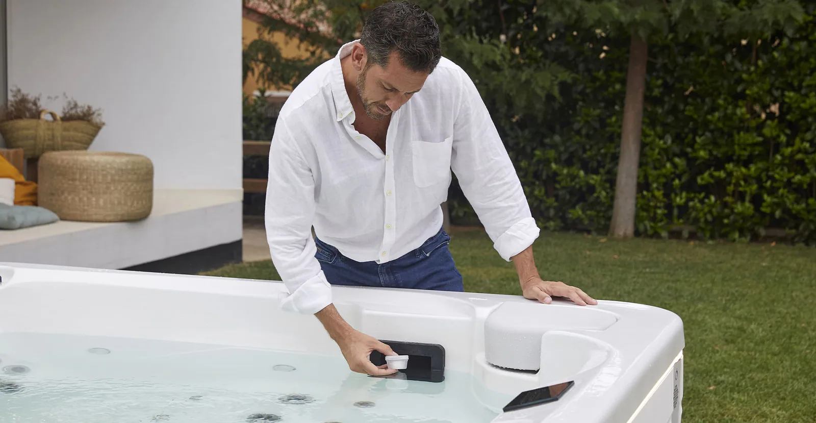 Hombre con camisa blanca sujetando una pieza junto al skimmer de un jacuzzi exterior en un jardín, imagen ilustrativa de reparación de jacuzzi en Bilbao