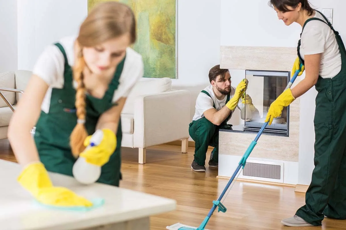 Tres operarios con petos verdes limpian un salón: uno friega el suelo, otro la chimenea y otra una mesa, imagen ilustrativa de una empresa de limpieza en Tenerife