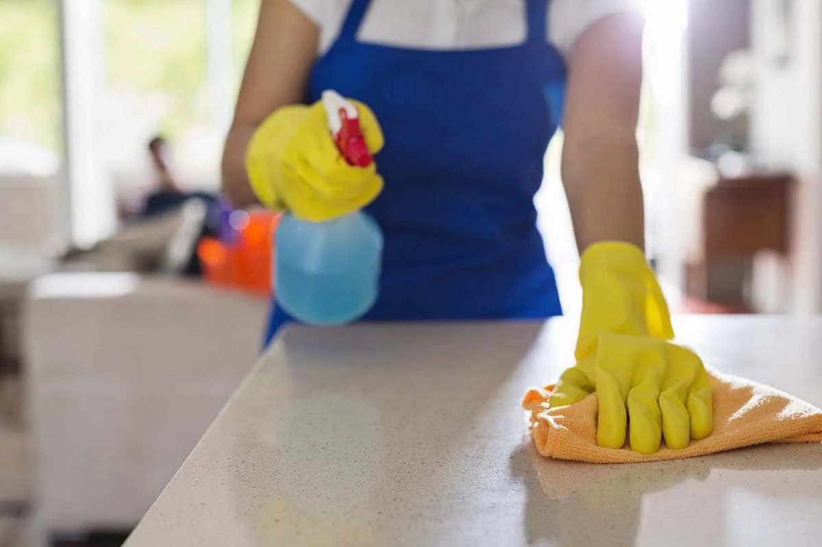 Manos con guantes amarillos pulverizan limpiador sobre la encimera de una cocina y la frotan con una bayeta naranja