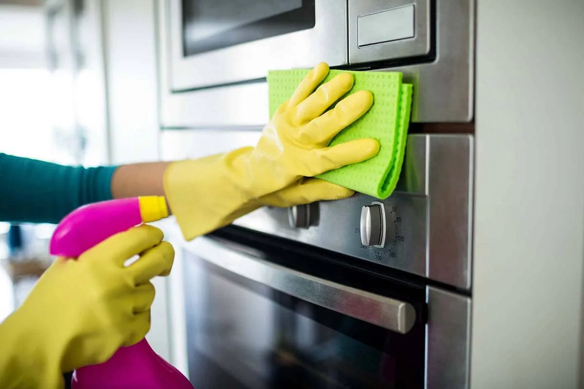 Mano con guante amarillo pasa una bayeta verde por el panel de acero de un horno empotrado, con un pulverizador rosa en la otra