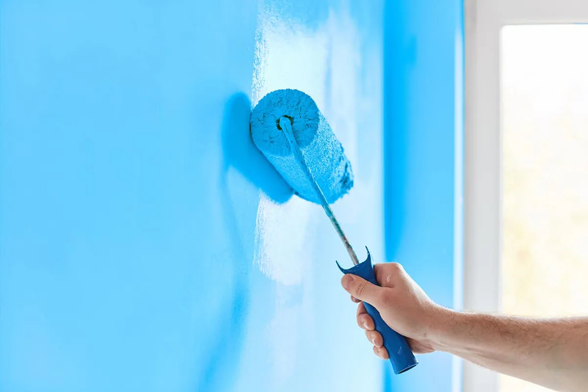 Mano con un rodillo extendiendo pintura azul celeste sobre una pared junto a una ventana, imagen ilustrativa para pintores en Tenerife
