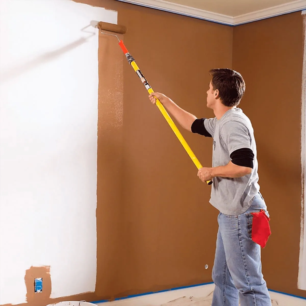 Operario con rodillo de mango extensible pintando de marrón una pared blanca, con cinta azul en el encuentro con el techo