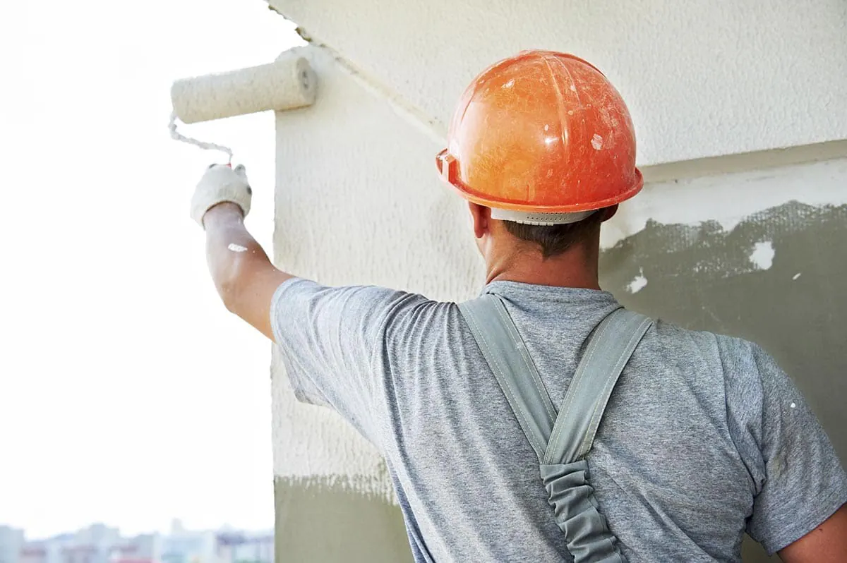 Operario con casco naranja y peto gris, de espaldas, pintando con rodillo un muro exterior en altura