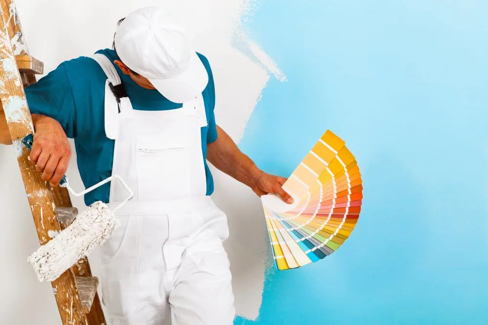 Pintor con peto blanco apoyado en una escalera de madera, con un rodillo en una mano y un abanico de colores en la otra