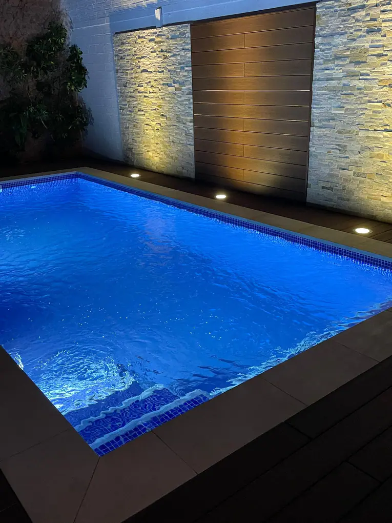 Piscina iluminada de noche con gresite azul, escalera interior y balizas empotradas en el pavimento junto a un muro de piedra