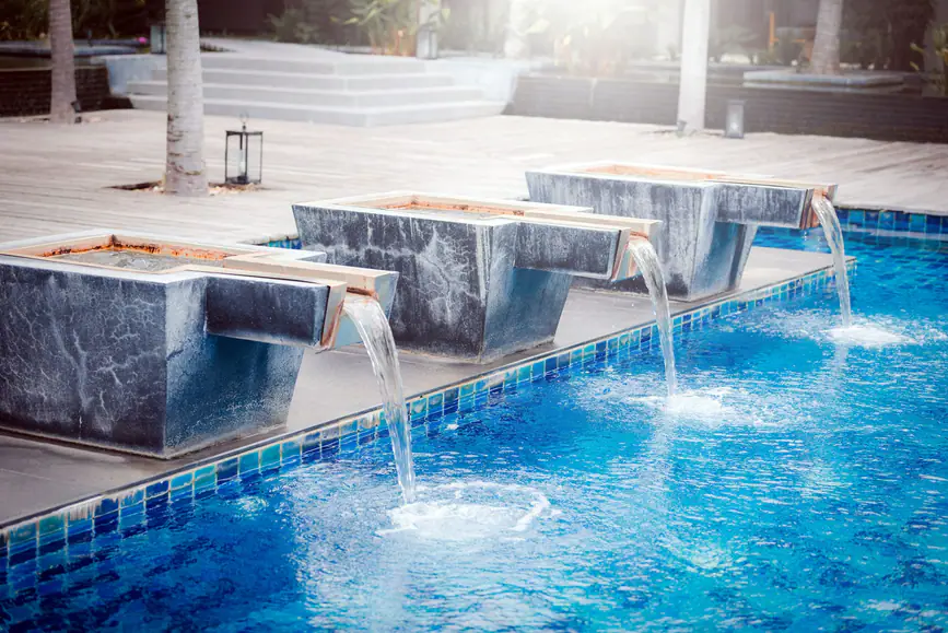 Tres caños de piedra oscura vertiendo agua sobre una piscina con gresite azul