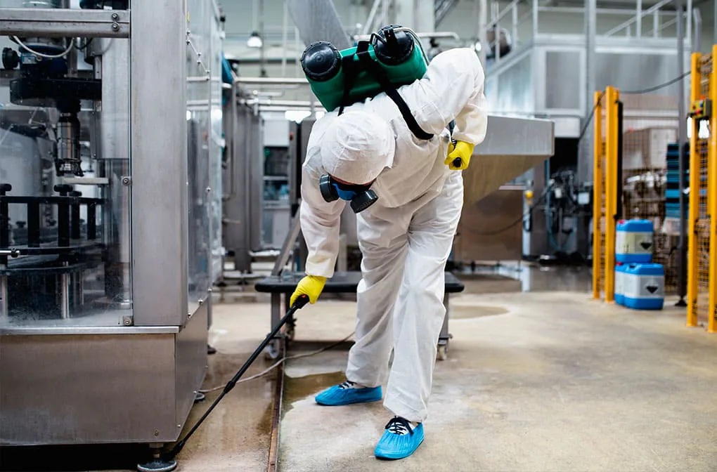 Técnico con mono blanco y fumigador de mochila rociando el suelo entre la maquinaria de una nave industrial