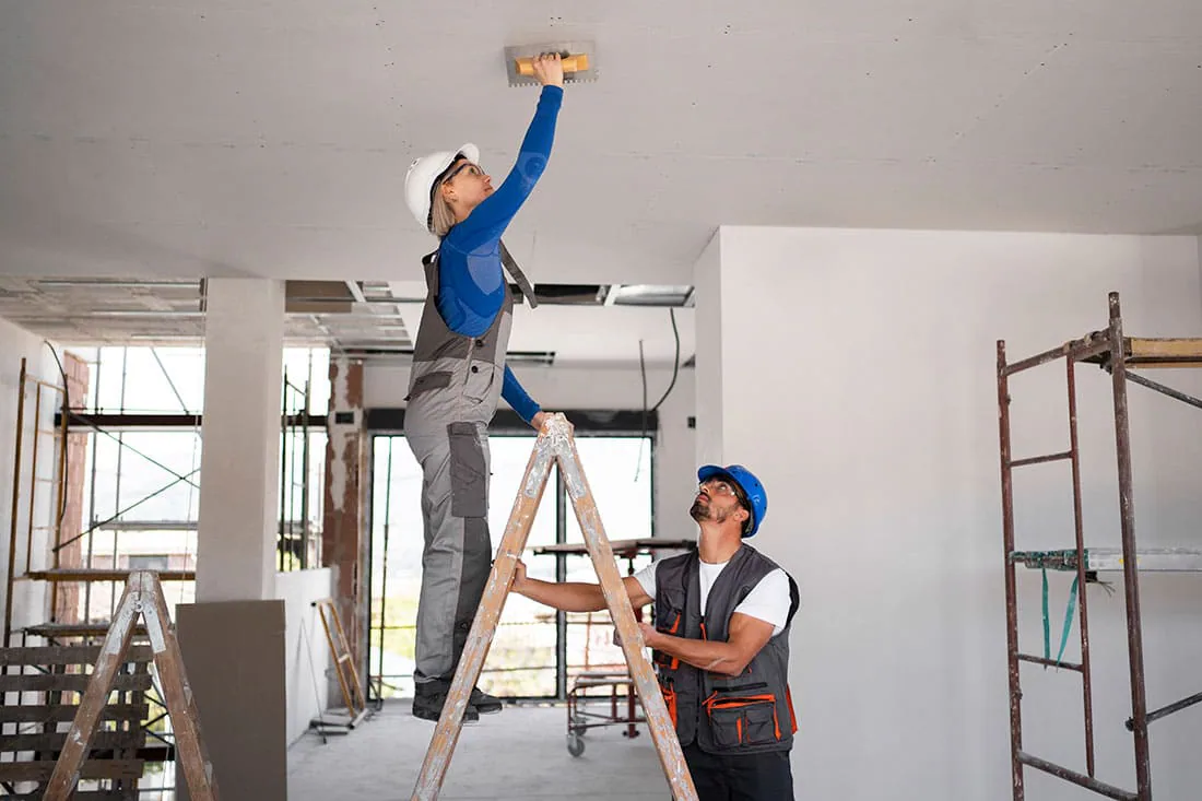 Operaria con casco alisando con llana el techo de una planta en obra mientras un compañero sujeta la escalera, imagen ilustrativa de reformas en Tenerife