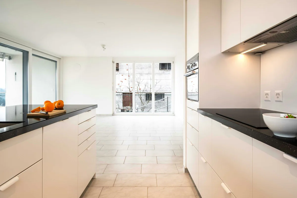 Cocina de muebles blancos y encimera negra abierta a un salón con suelo de baldosa clara