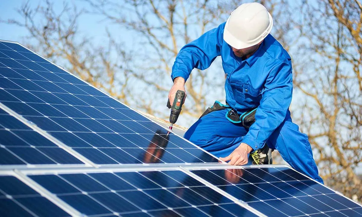 Instalación de placas solares: operario con casco blanco y mono azul fijando un módulo fotovoltaico con un taladro atornillador