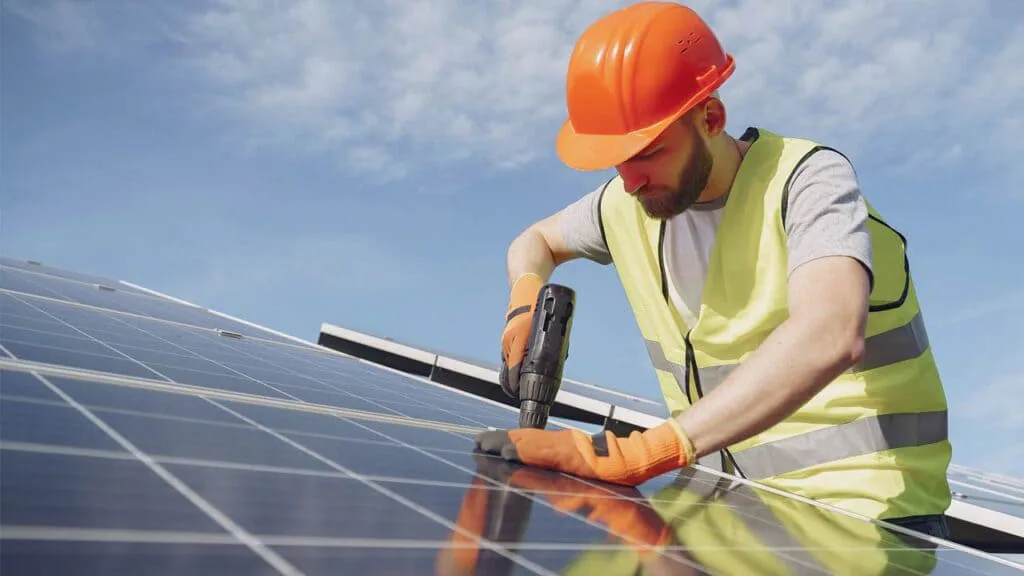 Instalador con casco naranja y chaleco reflectante atornillando con un taladro la fijación de un panel solar