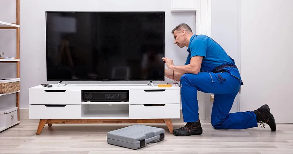 Técnico con mono azul arrodillado junto a un televisor, comprobándolo con las puntas de un polímetro