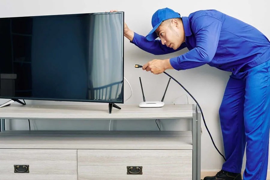 Técnico con gorra y mono azul conectando un cable detrás de un televisor, junto a un router