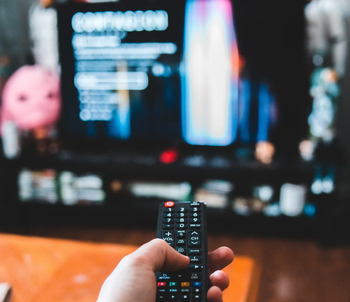 Mano sujetando un mando a distancia apuntando a un televisor encendido y desenfocado al fondo